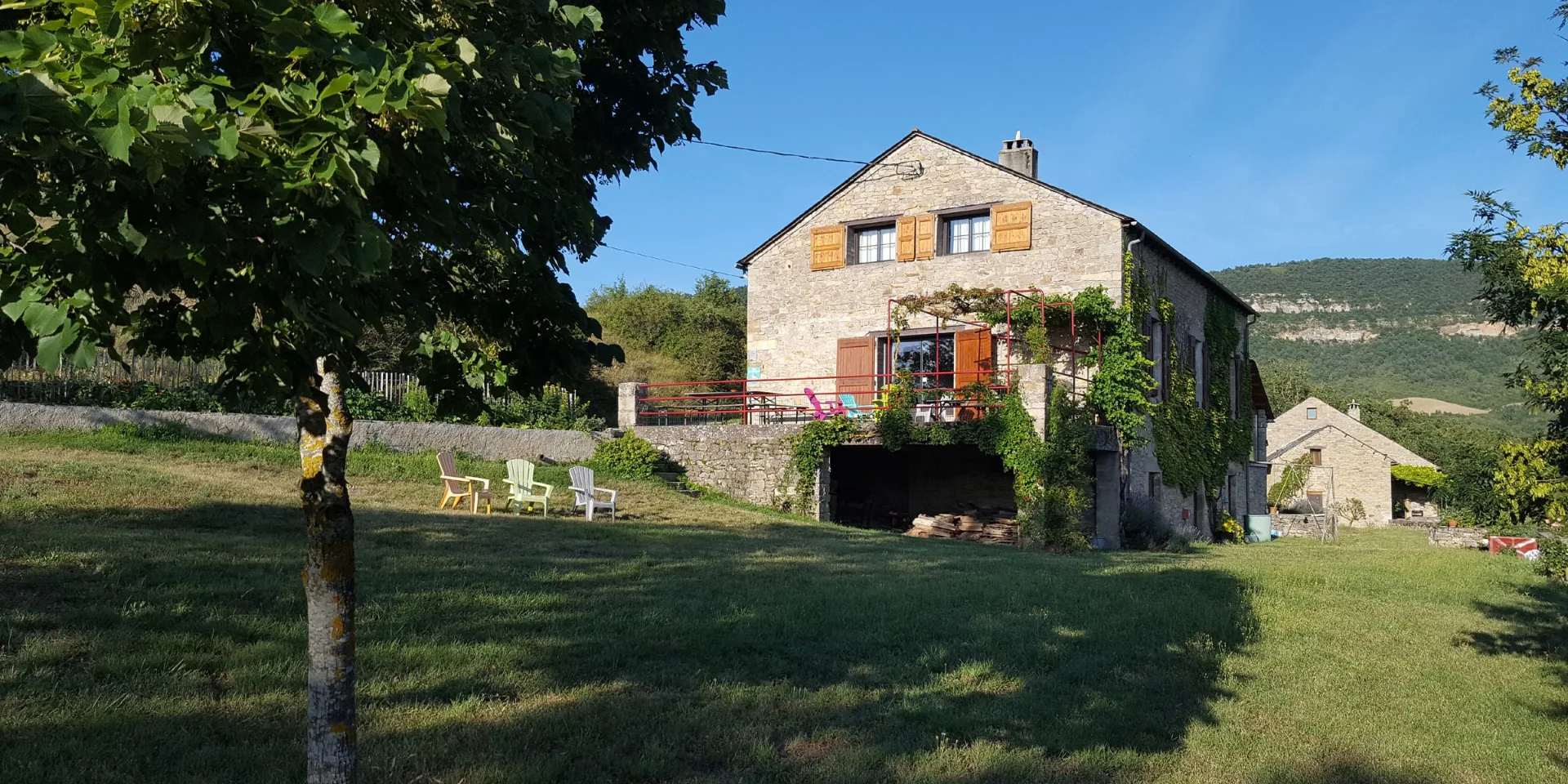 Gîte de groupe Gîte de Cabrières à Compeyre — Façade en pierre du gîte avec terrasse fleurie, mobilier de jardin et vue sur les collines, capacité 23 personnes