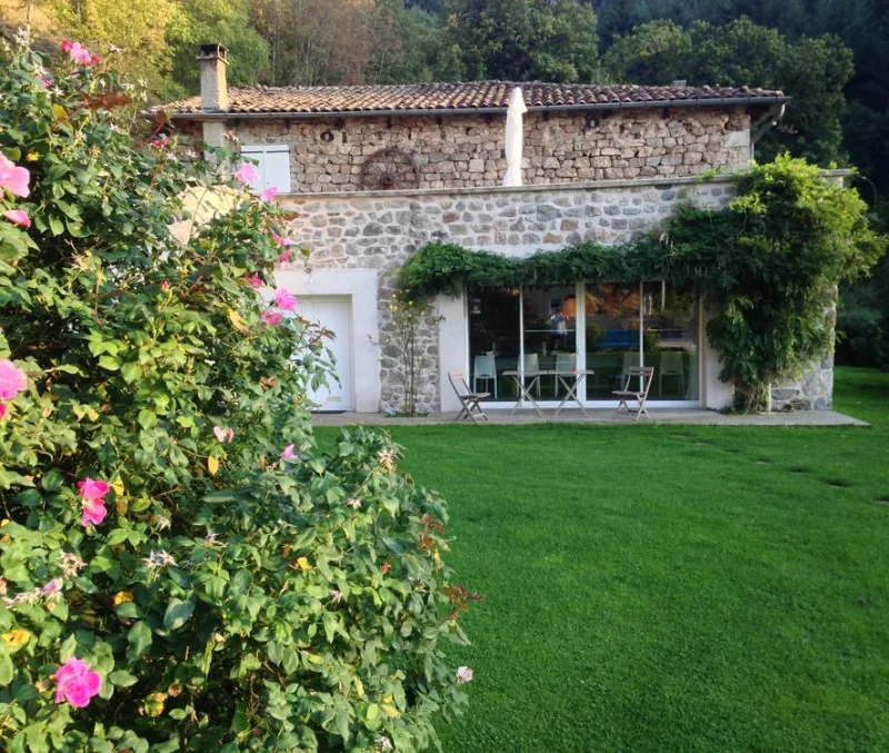 Terrasse repas ombragée devant les baies vitrées, façade en pierre couverte de vigne vierge