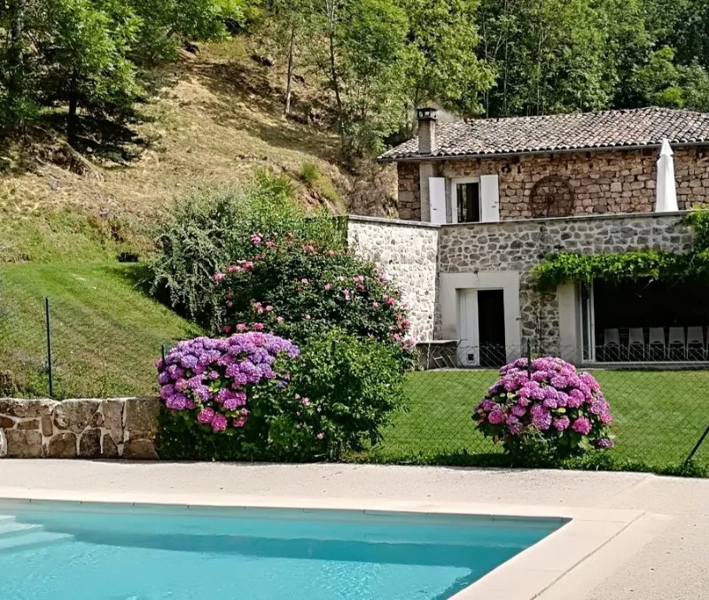 Façade en pierre du gîte vue depuis la piscine, entourée d'hortensias et de rosiers