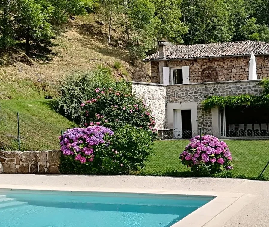 Gîte de groupe Les Combes en Ardèche à Accons — Façade en pierre du gîte vue depuis la piscine, entourée d'hortensias et de rosiers, capacité 17 personnes