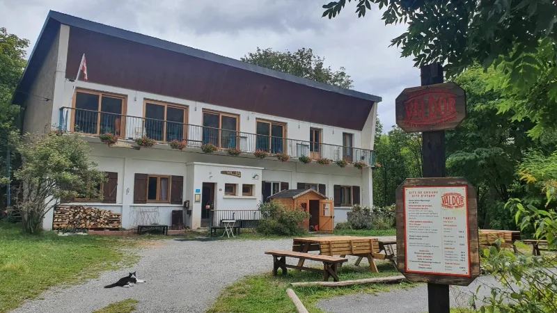 Façade blanche et toit orangé du gîte Le Val d'Or en été, fleurs aux balcons et chat sur le chemin