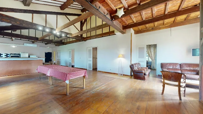 Grande salle de réception à poutres apparentes avec longue table et coin salon, idéale pour les groupes