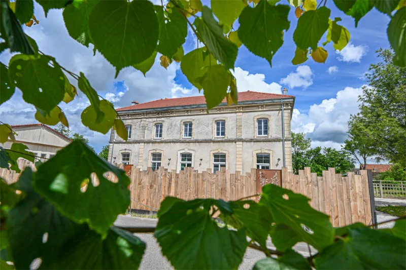 Façade en pierre de l'ancienne gare, entourée d'arbres, avec sa toiture en tuiles