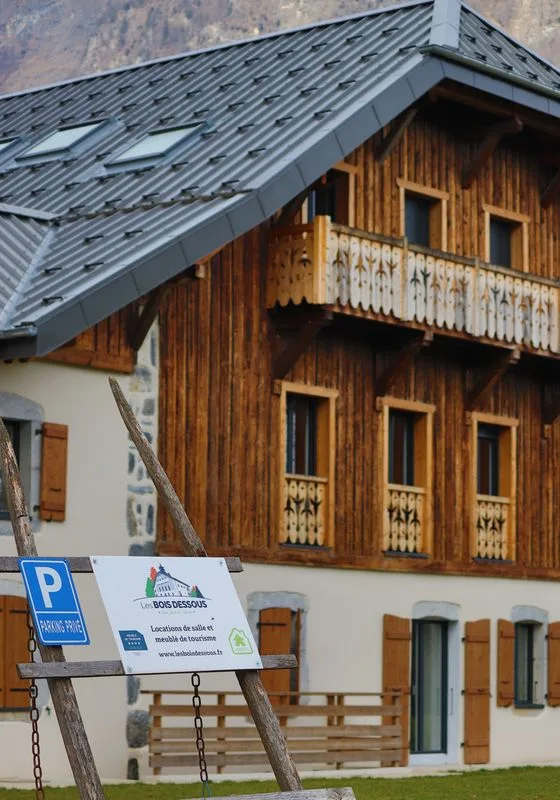 Façade en bois et pierre de la ferme savoyarde avec panneau Les Bois Dessous et parking