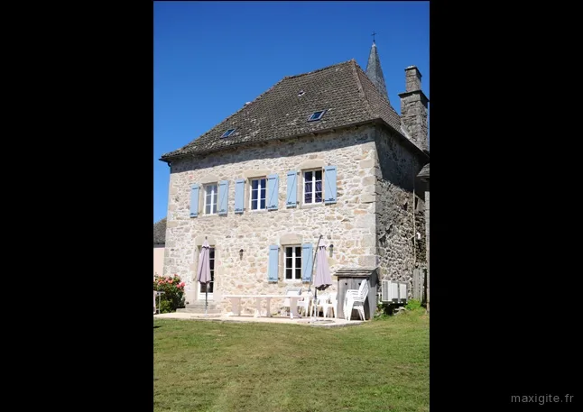 Façade en pierre avec volets bleus, terrasse aménagée avec table, chaises et jardin ombragé
