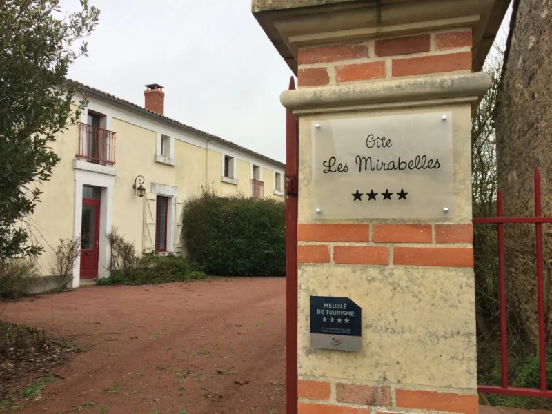 Façade principale du gîte avec panneau d'entrée, allée gravillonnée et volets rouges