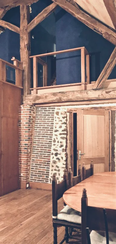 Grand salon sous charpente en bois avec mezzanine, murs en pierre et brique, table de repas ronde