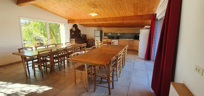 Grande salle à manger sous charpente en bois avec deux longues tables et cuisine ouverte équipée