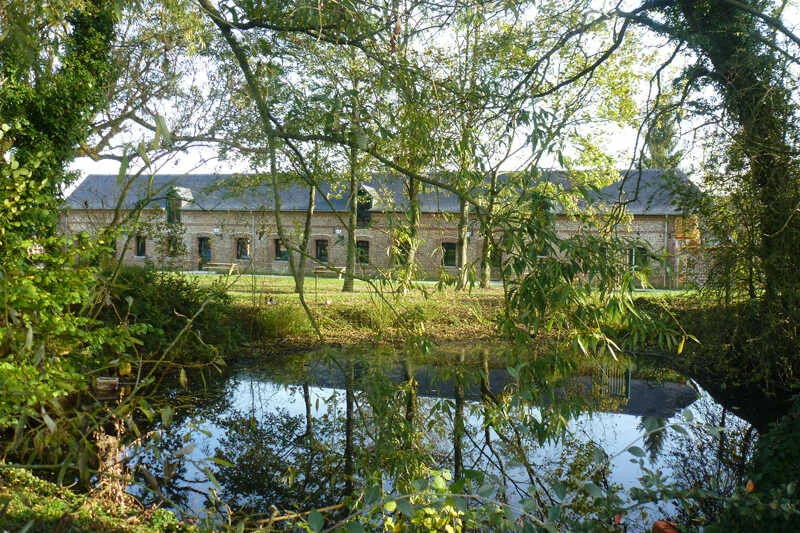 Étang bordé d'arbres avec vue sur la façade du gîte se reflétant dans l'eau