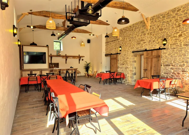 Grande salle commune en pierre avec nappes orange, bar et écran pour les soirées de groupe
