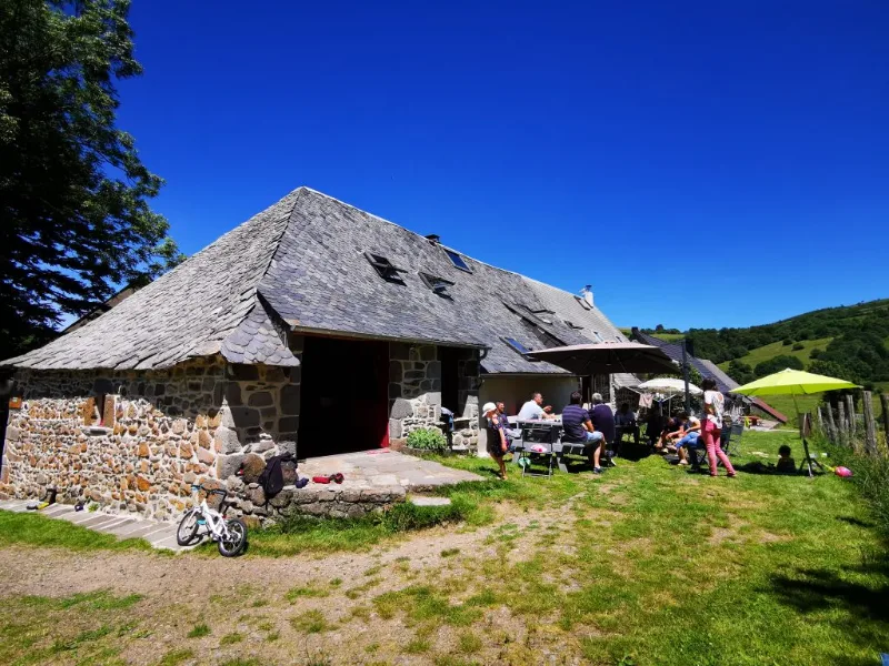 Façade en pierre du gîte avec toit de lauze et grande terrasse ombragée où un groupe partage un repas