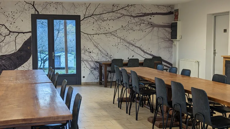 Salle a manger avec deux longues tables en bois, chaises noires et papier peint mural arbre