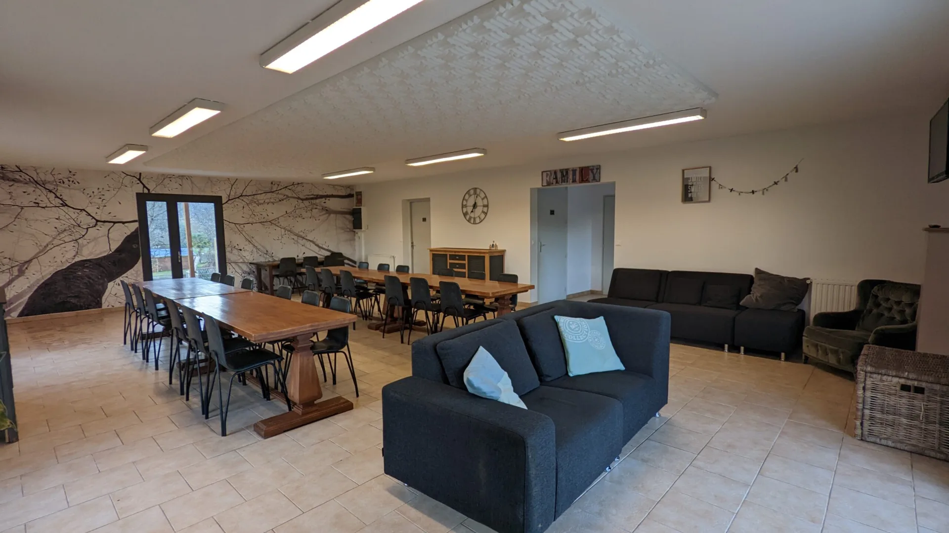 Grande piece de vie avec tables en bois pour 20 convives, canapes gris et decoration Family