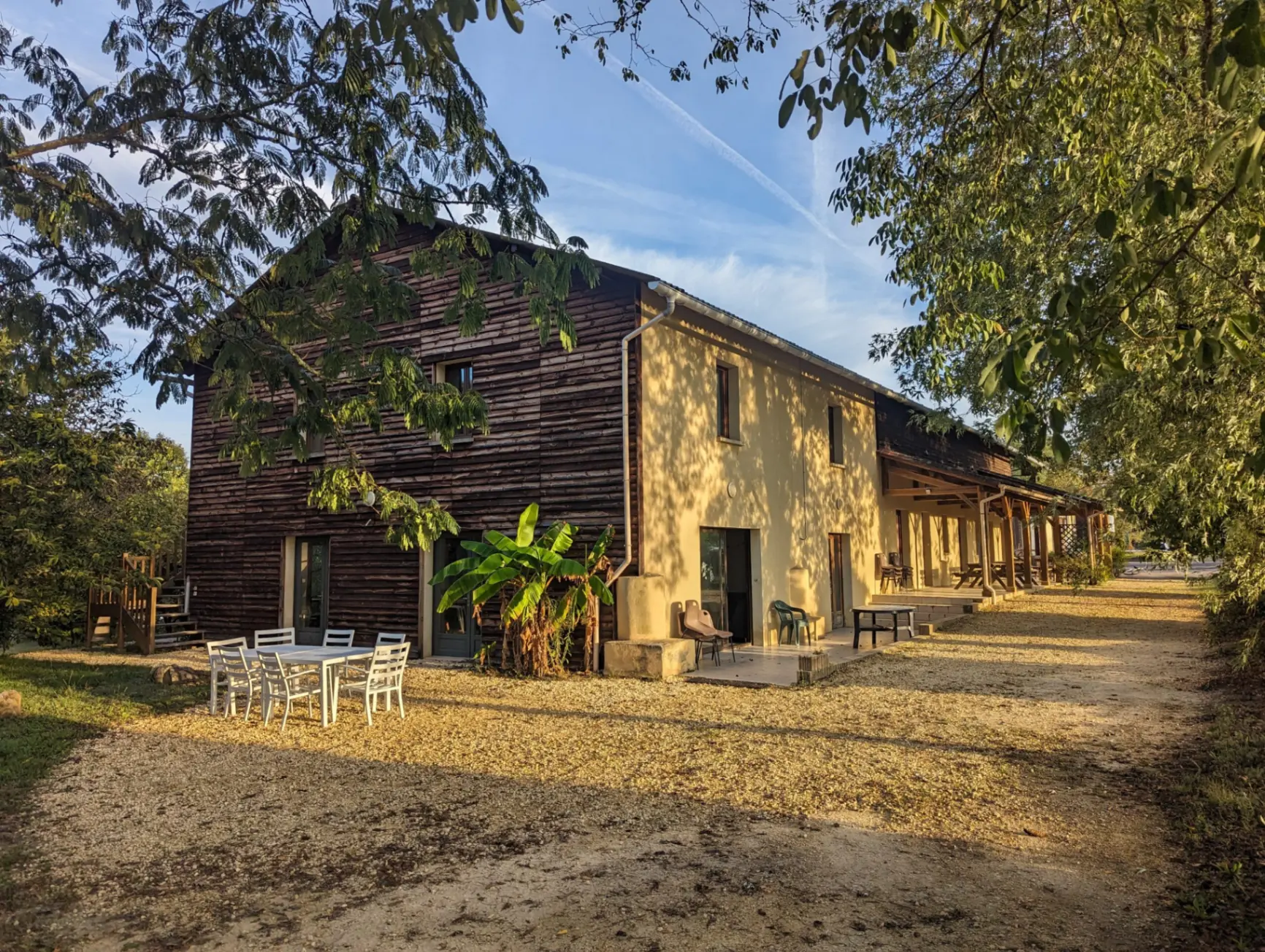 Gîte de groupe Gîte de Groupe aux 5 Châteaux à Vezac — Facade du gite en bardage bois et enduit avec terrasse, mobilier de jardin et bananier sous les arbres, capacité 20 personnes
