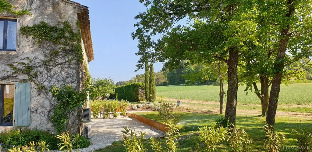 Façade latérale en pierre avec volets bleus, jardin fleuri et vue dégagée sur les champs environnants