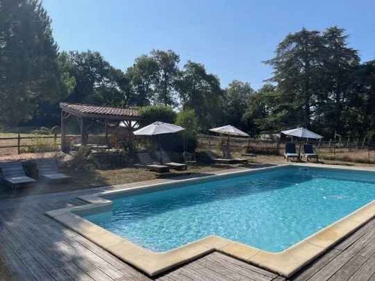 Piscine au sel avec terrasse en bois, transats, parasols blancs et pool house couvert entouré de grands arbres