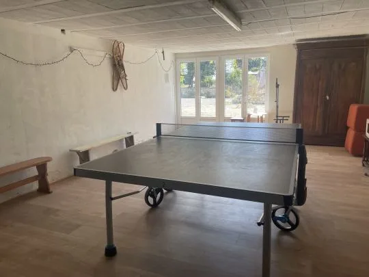 Salle de jeux intérieure avec table de ping-pong, parquet au sol, grande baie vitrée et armoire ancienne en bois