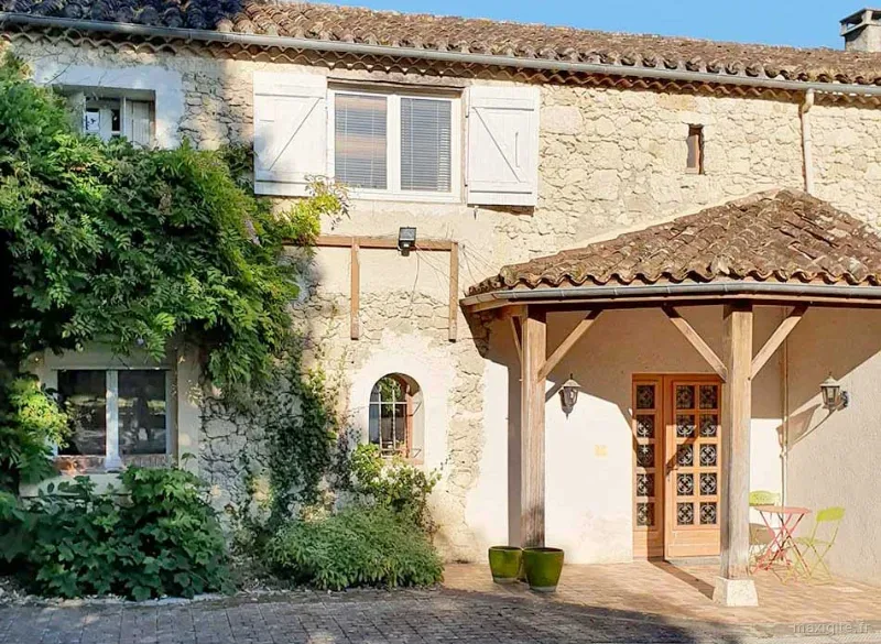 Façade en pierre du bâtiment principal avec porche couvert en bois, glycine grimpante, volets blancs et terrasse meublée