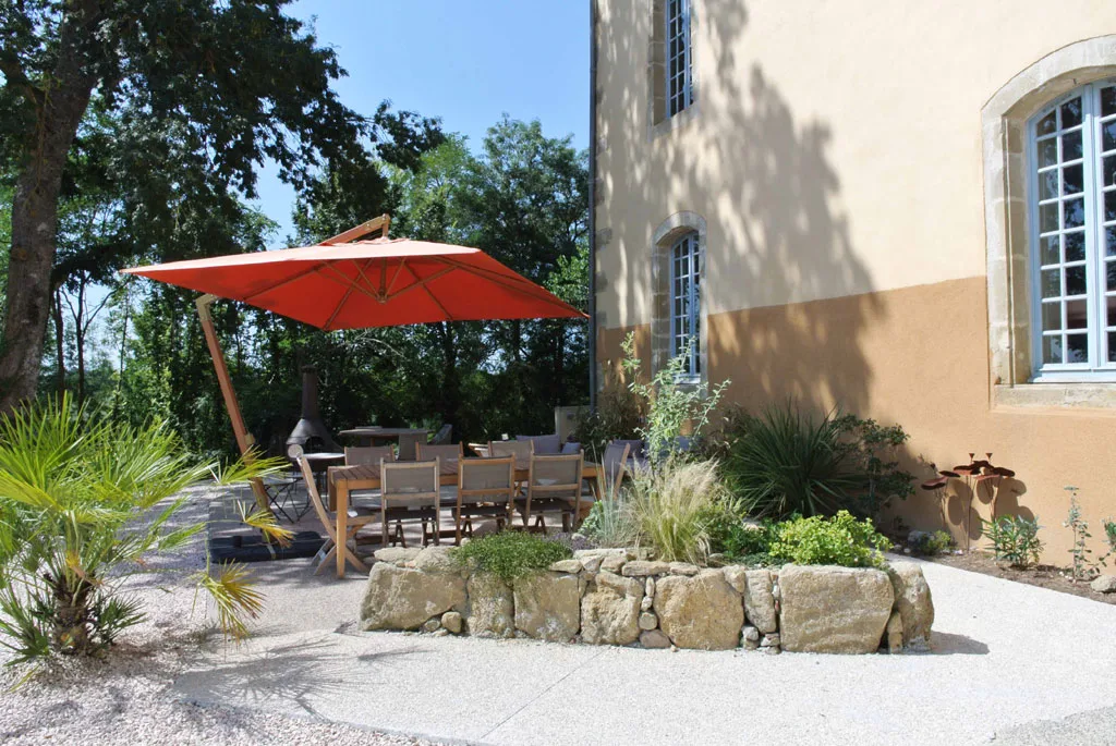 Terrasse extérieure avec parasol rouge, mobilier en bois et mur en pierre du château en arrière-plan