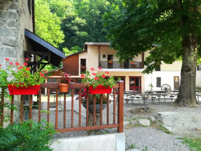Façade du gîte L'Escalusse avec bâtiment en pierre et toit brun, terrasse ombragée avec tables et chaises, clôture en bois rouge