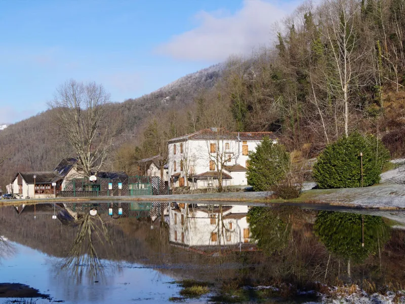 Bâtiments du domaine vus depuis le plan d'eau avec reflets, montagnes pyrénéennes enneigées en arrière-plan sous ciel bleu