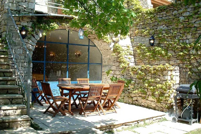 Terrasse ombragée avec table en bois et chaises devant la façade en pierre du mas provençal