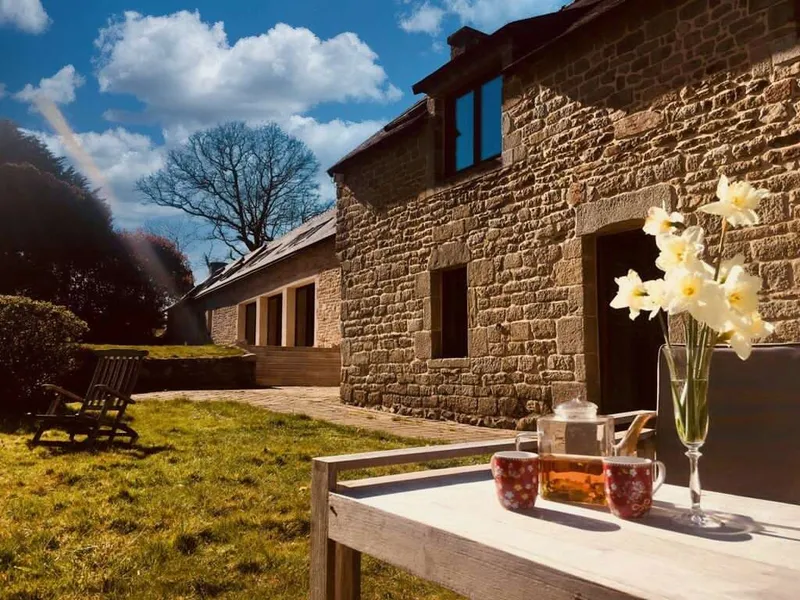 Façade en pierre du manoir sous un ciel bleu nuageux, terrasse en bois et bouquet de jonquilles au premier plan
