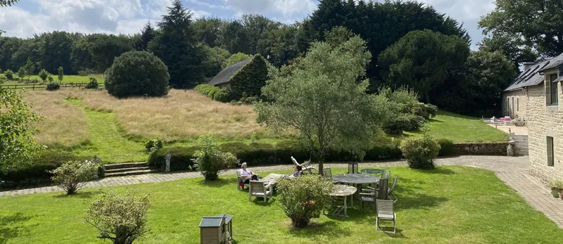 Grand jardin arboré avec coin salon extérieur sous un olivier et vue sur les dépendances en pierre