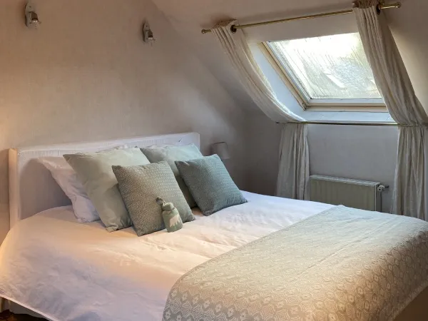 Chambre mansardée avec lit double, coussins vert d'eau, deux lampes de chevet allumées et velux vitré