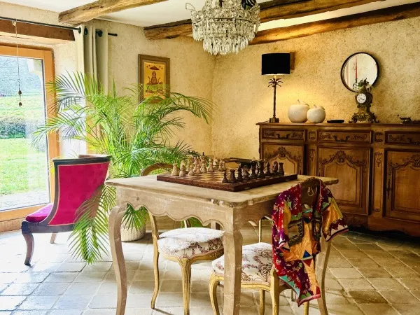 Salle à manger avec table en bois, échiquier, lustre en cristal, fauteuil rose et bibliothèque en fond