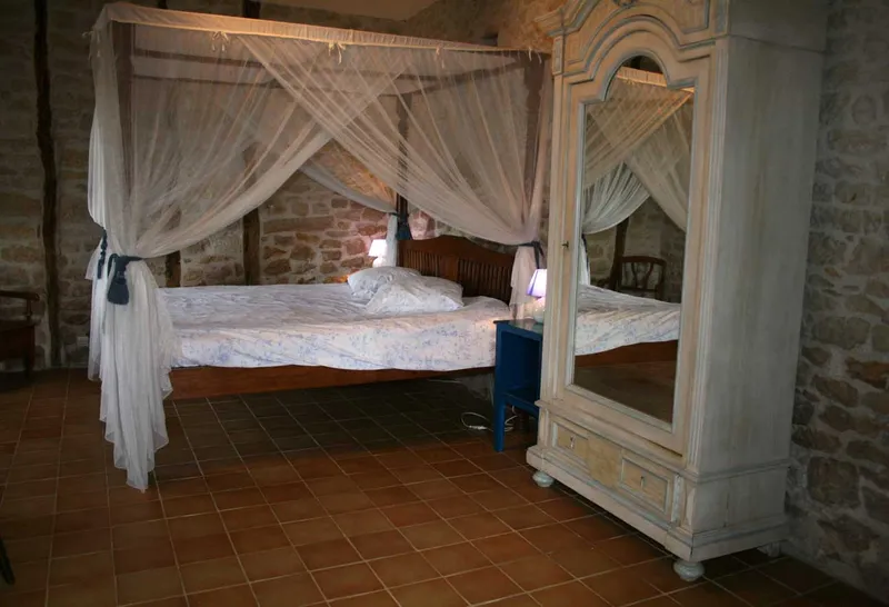 Chambre avec grand lit à baldaquin voilé, armoire ancienne à miroir et sol carrelé terre cuite