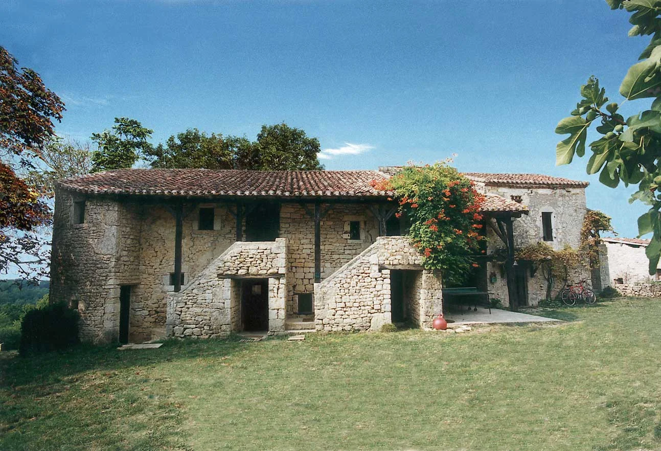 Gîte de groupe Le Hameau du Manoir de La Gabertie à Thézac — Façade en pierre du hameau avec toit de tuiles, cour engazonnée et vigne fleurie grimpant sur la pierre, capacité 28 personnes