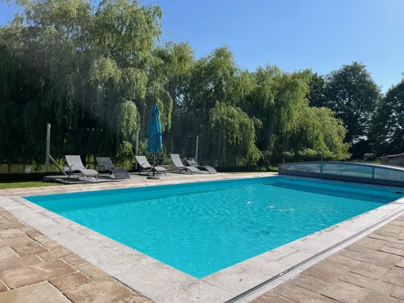 Piscine rectangulaire chauffée avec volet immergé, transats et parasol à l'ombre de grands saules pleureurs