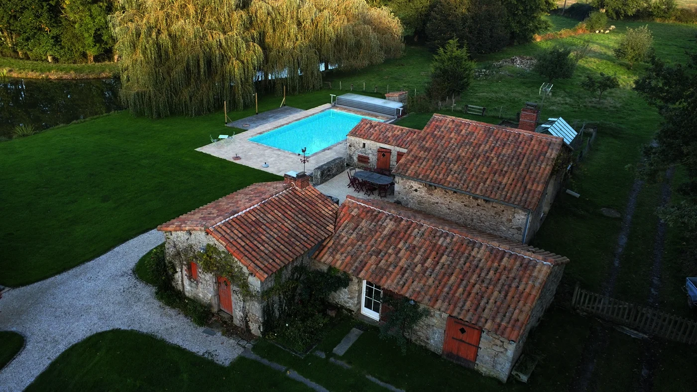 Gîte de groupe L'Étang Neuf à Sèvremont — Vue aérienne du corps de ferme en pierre, toits de tuiles rouges et piscine turquoise entourée de saules, capacité 20 personnes