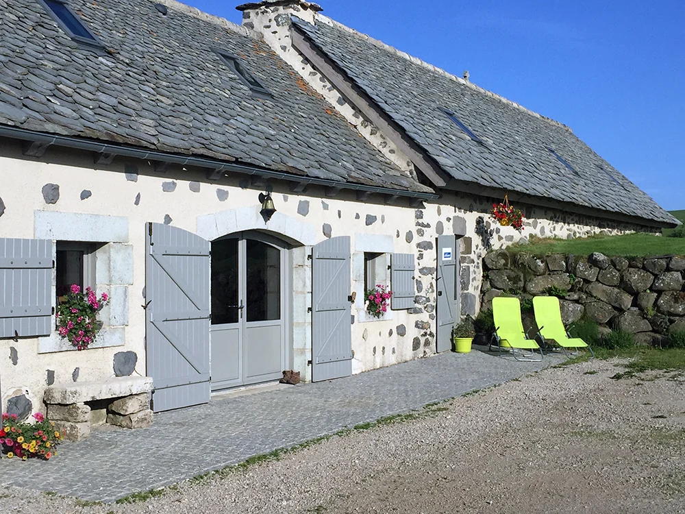 Gîte de groupe Le Buron de Veirières à Trizac — Façade en pierre volcanique du buron avec volets gris, toit de lauzes et terrasse gravillonnée, capacité 15 personnes