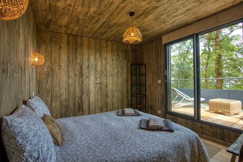 Chambre en bois avec lit double, linge fleuri gris et baie vitrée ouvrant sur une terrasse boisée