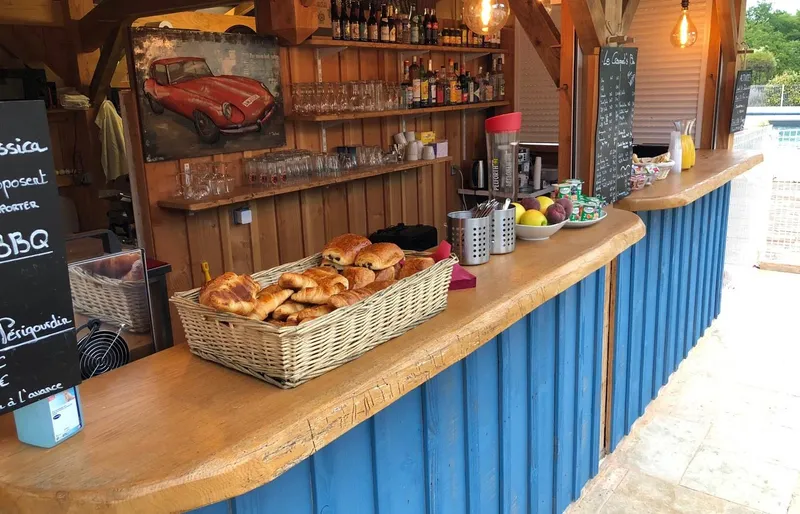 Bar extérieur en bois et bleu avec panier de viennoiseries, bouteilles en étagère et ardoise de menu au bord de la piscine