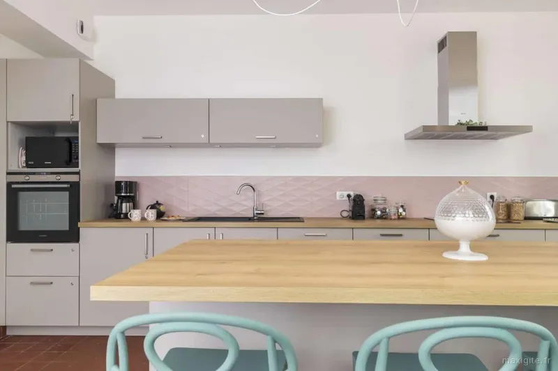 Cuisine équipée avec îlot en bois, hotte inox, crédence rose et chaises vert d'eau autour de la table