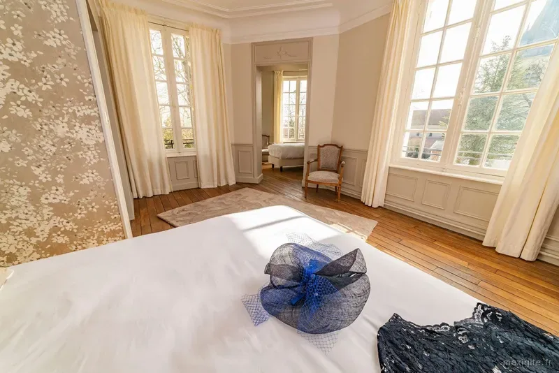 Chambre lumineuse avec grand lit blanc, parquet en bois, hautes fenêtres à petits carreaux et fauteuil ancien