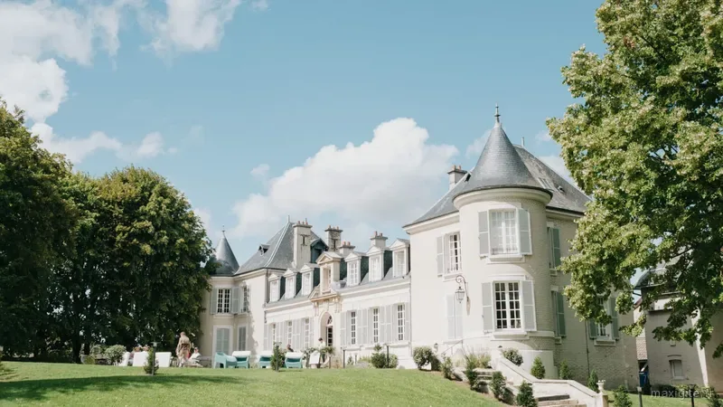 Façade du château avec tourelle en toit d'ardoise, volets gris-bleu et large pelouse verte entourée d'arbres