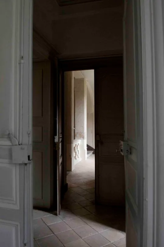 Enfilade de portes anciennes en bois ouvrant sur un couloir carrelé menant vers l'escalier de pierre voûté