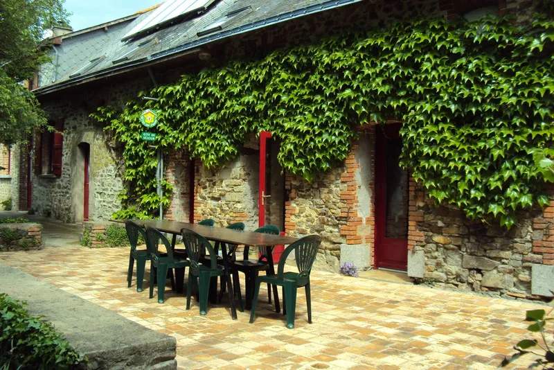 Façade en pierre ornée de lierre grimpant, portes rouges et grande table en bois pour les repas en plein air