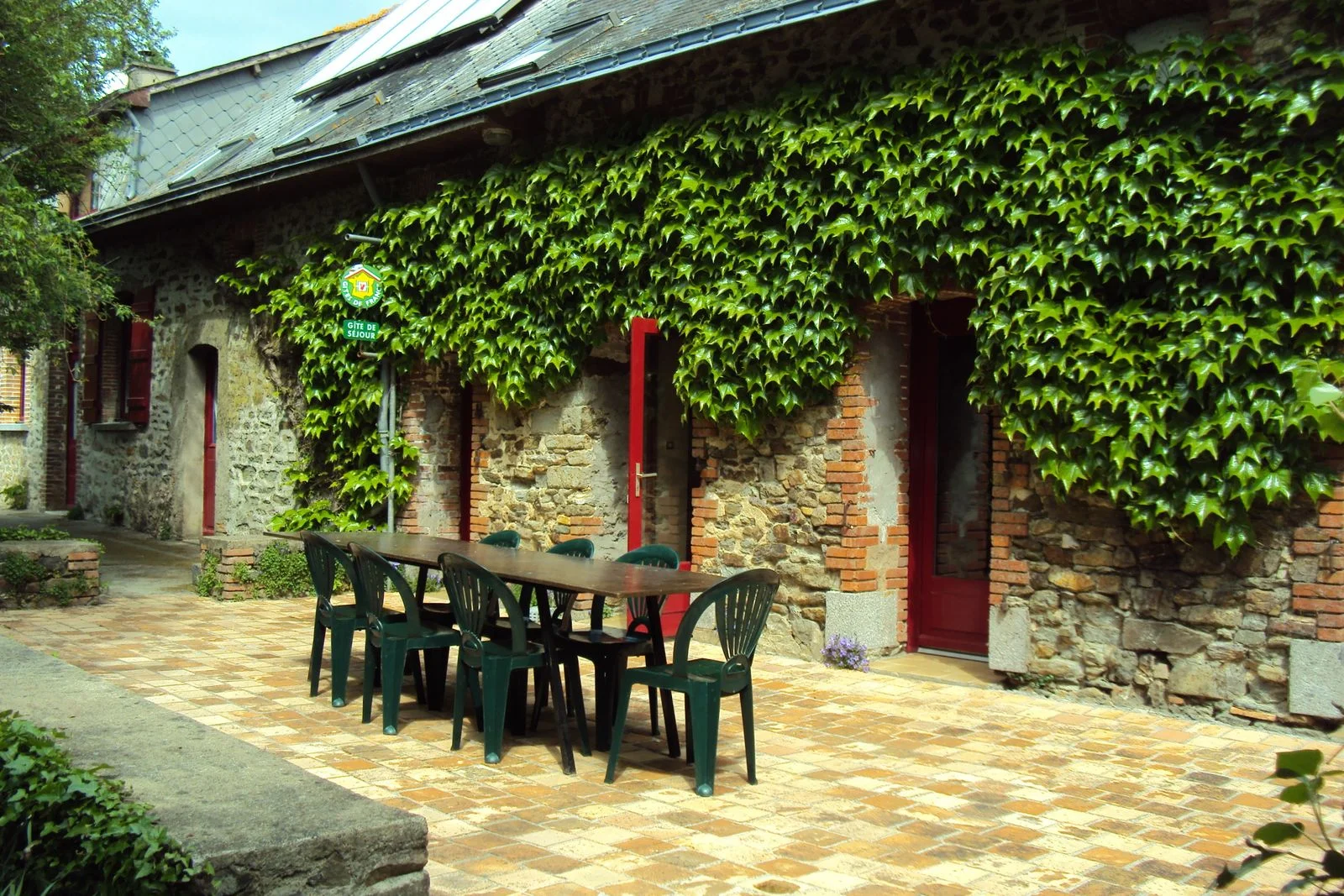 Gîte de groupe Les Feux Chauds à Bouchamps-lès-Craon — Façade en pierre ornée de lierre grimpant, portes rouges et grande table en bois pour les repas en plein air, capacité 15 personnes