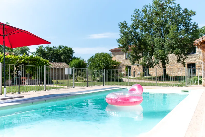 Façade en pierre du hameau avec piscine sécurisée, grand parasol rouge et jardin arboré