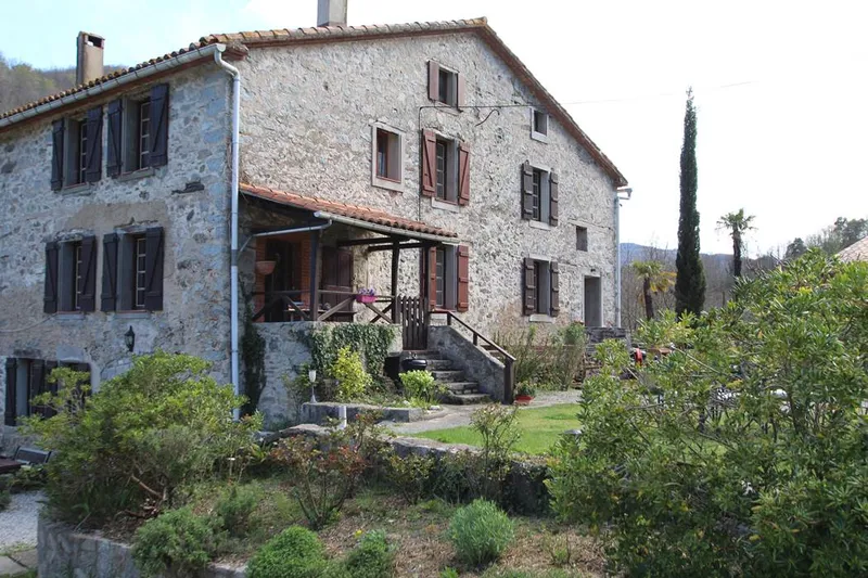 Façade en pierre du mas avec volets bruns, terrasse couverte et escalier de pierre menant au jardin