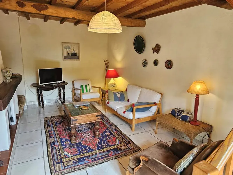 Salon cosy avec canapés clairs, poutres apparentes en bois, tapis persan et cheminée en pierre