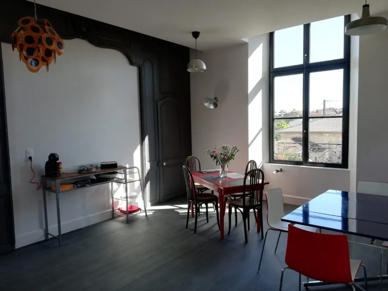 Salle à manger avec plusieurs tables en bois colorées et chaises dépareillées, sous une ancienne cheminée noire moulurée