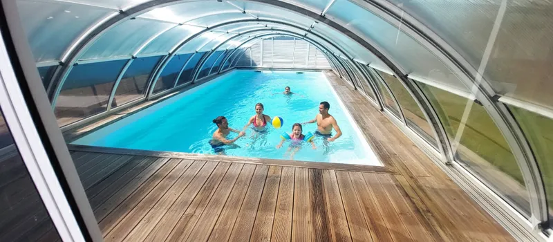 Piscine couverte sous abri translucide en arc, terrasse en bois et grandes baies vitrées laissant entrer la lumière