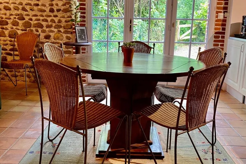 Salle à manger avec grande table ronde en bois et chaises en osier, cuisine ouverte en arrière-plan