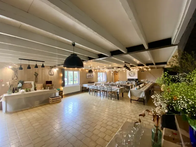 Grande salle commune aux poutres blanches avec longues tables dressées et coin cuisine en fond